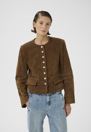 Žena s kratkom kosom, u smeđoj antilop jakni na kopčanje s preklopnim džepovima i širokim svijetloplavim trapericama, stoji na jednobojnoj pozadini.