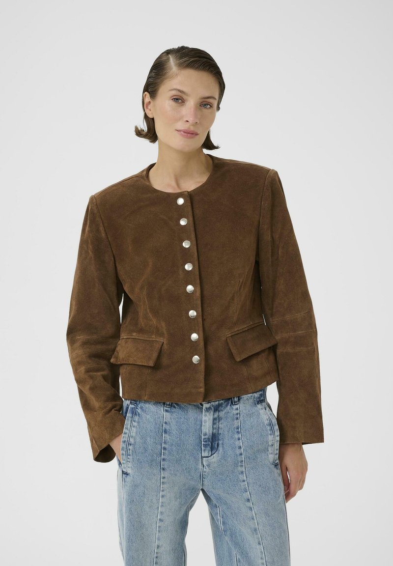 Mujer de cabello corto con chaqueta marrón de ante abotonada con bolsillos de solapa y vaqueros anchos azul claro, posando frente a un fondo liso.