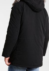 Parka noire avec capuche, ornée d'une bordure en fausse fourrure, manches longues et coupe décontractée. Texture lisse avec détails de coutures visibles.