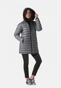 Manteau en duvet gris avec une capuche, présentant un motif matelassé, une fermeture éclair à l'avant et des poches latérales. Associé à un jean noir et des baskets.