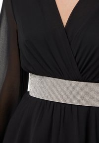 Abito nero con scollatura a V profonda, maniche lunghe trasparenti e una cintura decorata con strass argentati in vita, creando una silhouette aderente.