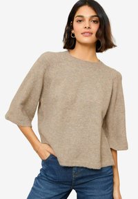 Hellbrauner Strickpullover, Rundhals, kurze Raglanärmel, lockere Passform, strukturierter Stoff, kombiniert mit blauen Jeans.
