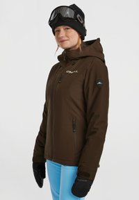 Bruine waterdichte skijas met een capuchon, ritszakken en geborduurd logo. Draagt met blauwe broeken en zwarte handschoenen, aangevuld met skibril.