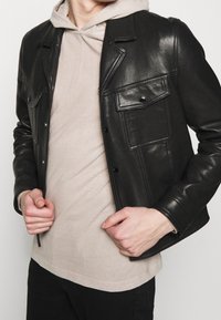 Person som bär en beige hoodie och en svart läderjacka med fickor, håller i jackans kanter med båda händerna.