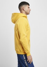 Felpa gialla con cappuccio, tasca a marsupio, polsini e orlo a coste. Modella indossa pantaloni sportivi blu con una striscia di logo lungo il lato.