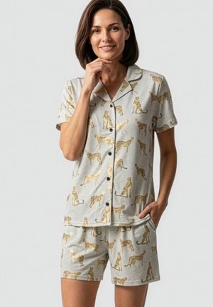Vrouw draagt lichtgrijze korte mouw blouse met knopen en korte broek met gouden luipaardprint, glimlacht met één hand op de kin en één in de zak.