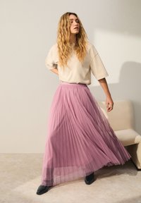 Jupe longue plissée rose transparent avec une taille élastique, associée à un t-shirt beige ample et des bottines noires à bout carré.