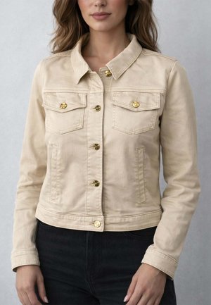 Donna che indossa una giacca beige con bottoni dorati e tasche sul petto, abbinata a pantaloni neri, su uno sfondo grigio chiaro uniforme.