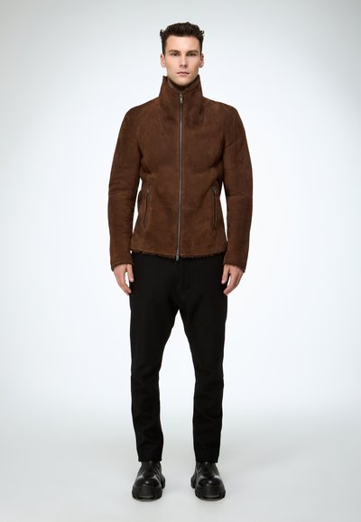 Braune Wildlederjacke mit hohem Kragen und Frontreißverschluss, mit zwei seitlichen Reißverschlusstaschen. Dazu schwarze, schmal zulaufende Hose und klobige Schuhe.