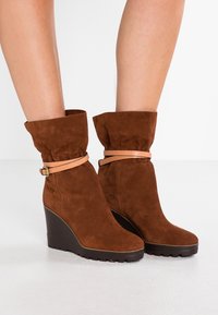 Bruna mockakängor i kilklack med rullad krage och ett lätt läderband runt ankeln, med en strukturerad sula för bra grepp.