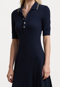 Marineblå ribbad klänning med korta ärmar, polokrage, guldfärgade knappar och två guldstripes på kragen. Utsvängd kjoldesign.