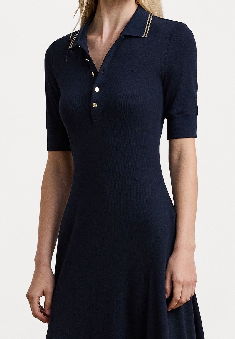 Marineblå ribbad klänning med korta ärmar, polokrage, guldfärgade knappar och två guldstripes på kragen. Utsvängd kjoldesign.