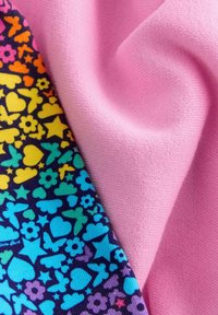 Un tessuto colorato presenta uno sfondo scuro con motivi di cuori, stelle e farfalle in tonalità vivaci, affiancato da un tessuto rosa chiaro e liscio.