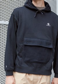 Uomo che indossa una felpa nera con cappuccio Converse con tasca frontale e pantaloni beige, in piedi di fronte a uno sfondo in rete metallica.