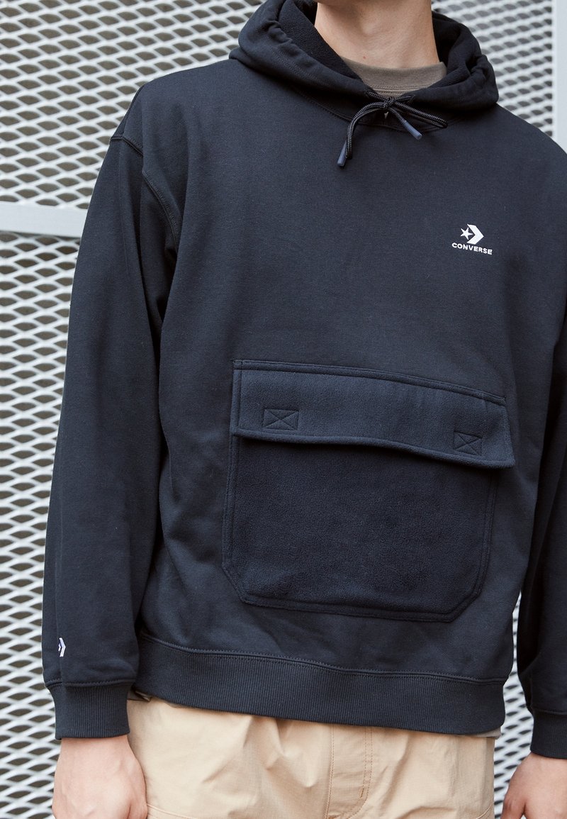 Uomo che indossa una felpa nera con cappuccio Converse con tasca frontale e pantaloni beige, in piedi di fronte a uno sfondo in rete metallica.