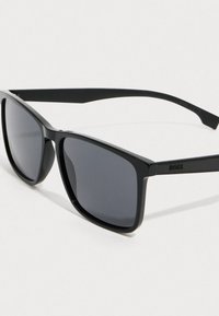 Mustad mustad päikeseprillid tumedate läätsedega ja kitsaste kätega, mille templites on BOSS logo valge tausta vastu.