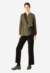 Olivově zelená košile s dlouhým rukávem, černé kalhoty a zelené slip-on boty. Černá tkaná crossbody taška s nastavitelným popruhem. Jednoduchý, funkční design.