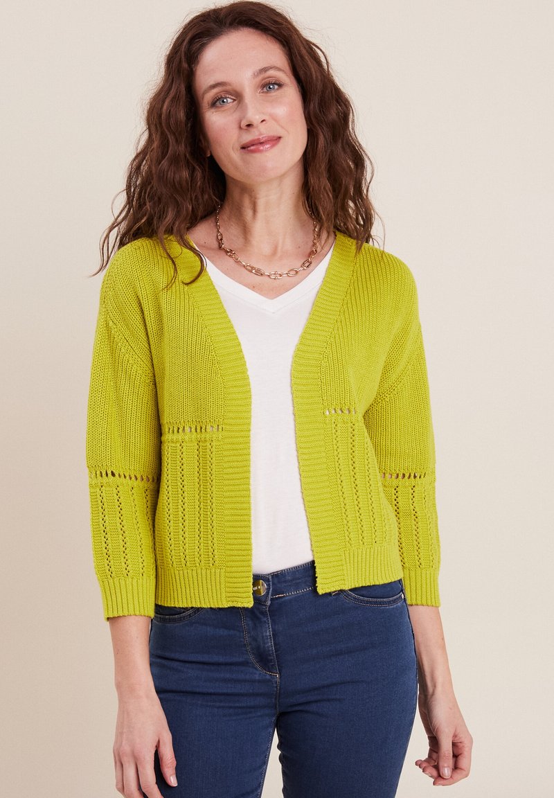 Cardigan a maglia verde lime con maniche a tre quarti, caratterizzato da dettagli in lavorazione a giorno e una vestibilità rilassata, indossato sopra una maglietta bianca a scollo a V.