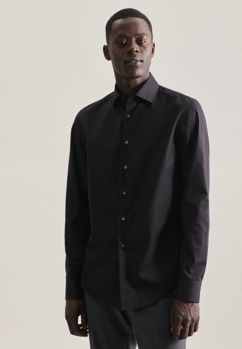 Chemise noire à manches longues, devant boutonné, en tissu lisse, coupe ajustée, ourlet courbé, col classique et accents de boutons discrets.