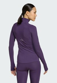 Haut de sport long violet avec col montant, design ajusté et passe-pouces aux poignets ; comporte un détail froncé au dos.