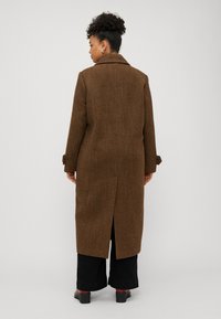 Lungo cappotto a fantasia houndstooth marrone con colletto a scollatura, spacco posteriore e polsini arrotolati. Indossato sopra pantaloni neri a gamba larga e scarpe con tacco.