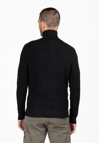 Maglione nero a collo alto con texture a coste e polsini aderenti, abbinato a pantaloni cargo verde oliva. La vista posteriore mostra un design senza cuciture.