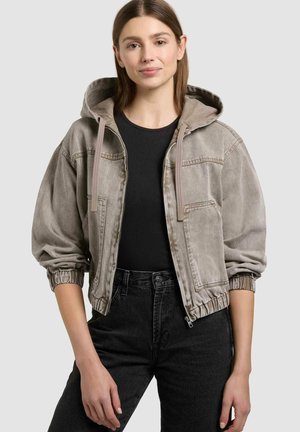 Tânără purtând o jachetă cu glugă gri, largă, cu fermoar peste un tricou negru și blugi negri cu talie înaltă, stând în picioare pe un fundal simplu.