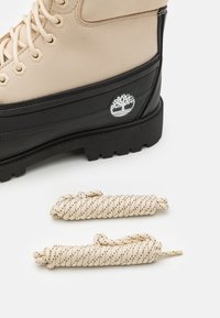 Botín de cuero beige con cordones y suela de goma negra, con un logo de árbol plateado, acompañado de cordones crema con motas negras.