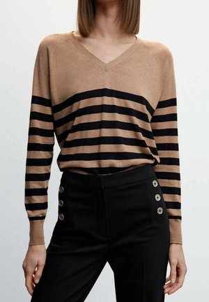 Femme portant un pull col en V beige avec des rayures horizontales noires et un pantalon noir taille haute avec des boutons décoratifs sur les hanches.