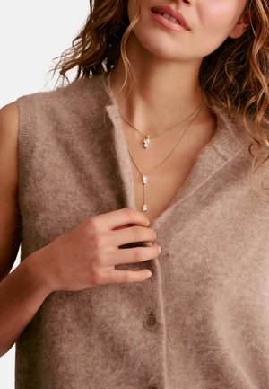 Frau trägt eine flauschige beige ärmellose Strickjacke und berührt den Halsausschnitt. Sie trägt zwei goldene Schichtenketten mit klaren Edelsteinanhängern.