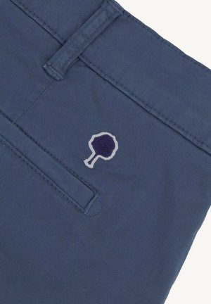 Tissu bleu marine avec passants de ceinture cousus, couture de poche horizontale, et petit logo d'arbre brodé en fils violet foncé et clair.