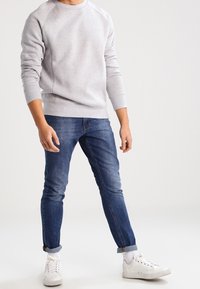Persona che indossa un maglione grigio chiaro, jeans blu attillati con orli, calzini bianchi e sneaker bianche, in piedi davanti a uno sfondo neutro.