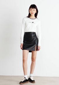 Falda de imitación de cuero negra con una abertura lateral, combinada con una camisa blanca de manga larga que presenta un logo negro y un cuello acanalado. Zapatillas negras.