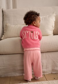 Maglione rosa lavorato a maglia con scritta "SMILE" e dettaglio a cuore, abbinato a pantaloni a coste rosa chiaro con orli arricciati, su sfondo neutro.