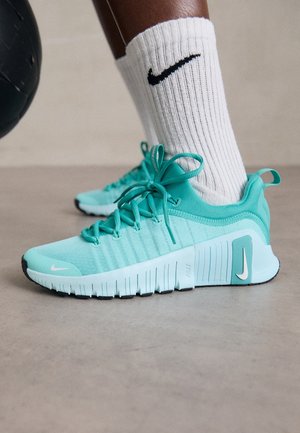 Pied portant une chaussette Nike blanche et une chaussure de course Nike turquoise, debout sur un sol lisse à côté d’un ballon médicinal noir.