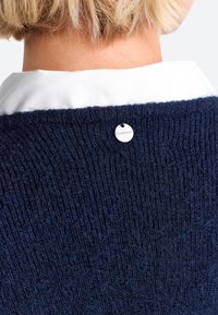 Maglione lavorato a maglia blu navy con una superficie a coste e una camicia bianca con collo visibile sotto. Caratterizzato da un piccolo tag argentato sul retro.