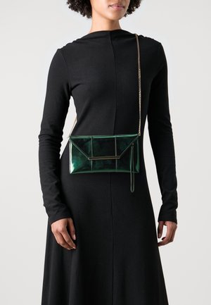 Femme portant une robe noire à manches longues et un petit sac bandoulière vert foncé brillant avec une chaîne dorée et un détail en barre métallique.