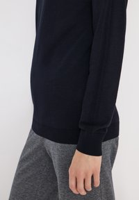 Personne portant un pull noir à manches longues et un pantalon gris texturé, le bras détendu le long du corps, sur un fond blanc uni.