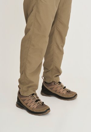 Benen met lichtbruine broek en bruine wandelschoenen met zwarte zolen en veters met rode accenten op een witte achtergrond.