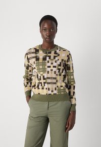 Pull en tricot avec un motif géométrique en vert, beige et marron, doté d'un col rond et de poignets et d'ourlet côtelés, associé à un pantalon vert.