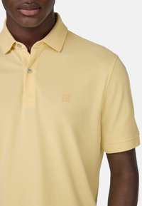 Geltonas polo marškinėlis, pagamintas iš tekstūruotos medžiagos. Savybės: apykaklė, trijų mygtukų užsegimas ir subtilus logotipo akcentas krūtinėje. Trumpomis rankovėmis.
