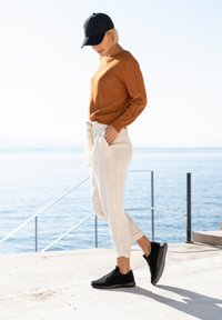 Brown knitted sweater, white textured pants tied at the waist, and black sneakers. Background features water and a clear sky.