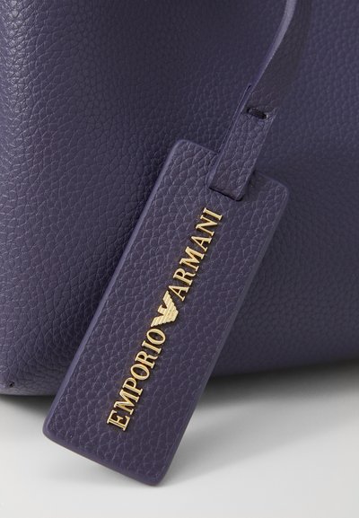 Geantă din piele de culoare violet închis, cu o etichetă aurie embosată pe care este afișat proeminent "EMPORIO ARMANI".