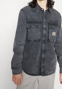 Šedá džínová košile se dvěma náprsními kapsami, zapínáním na knoflíky a sklopeným límcem. Má malou nášivku loga na levé kapse.