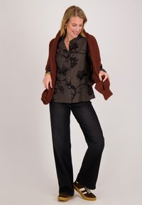 Vrouw in een zwarte bloes met bloemenprint, donkere wijde broek, bruine sneakers en een bruine gebreide trui over haar schouders gedrapeerd.