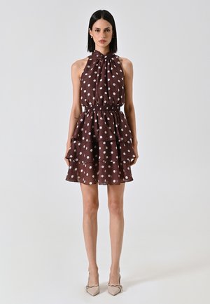 Robe à pois bruns avec un col halter, des volants et une taille élastique. Fabriquée en tissu léger. Présente des motifs circulaires blancs.