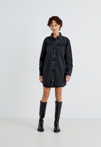 Vestido campero de mezclilla negra con mangas largas, cierre de botones, dos bolsillos en el pecho y un corte relajado, combinado con botas negras hasta la rodilla.