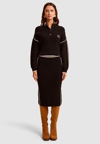 Vestido de punto negro acanalado con cuello tipo camisa, rayas crema contrastantes y una silueta ajustada al cuerpo. Botas marrones hasta la rodilla.