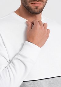 Weißes und graues Sweatshirt mit gerippten Bündchen. Verfügt über einen horizontalen grauen Streifen auf Taillenhöhe und hat eine strukturierte Stoffoberfläche.