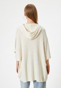 Poncho con capucha tejido en color beige claro, con textura acanalada; mangas cortas y dobladillo en forma de concha. La vista trasera muestra la capucha y el patrón diagonal.